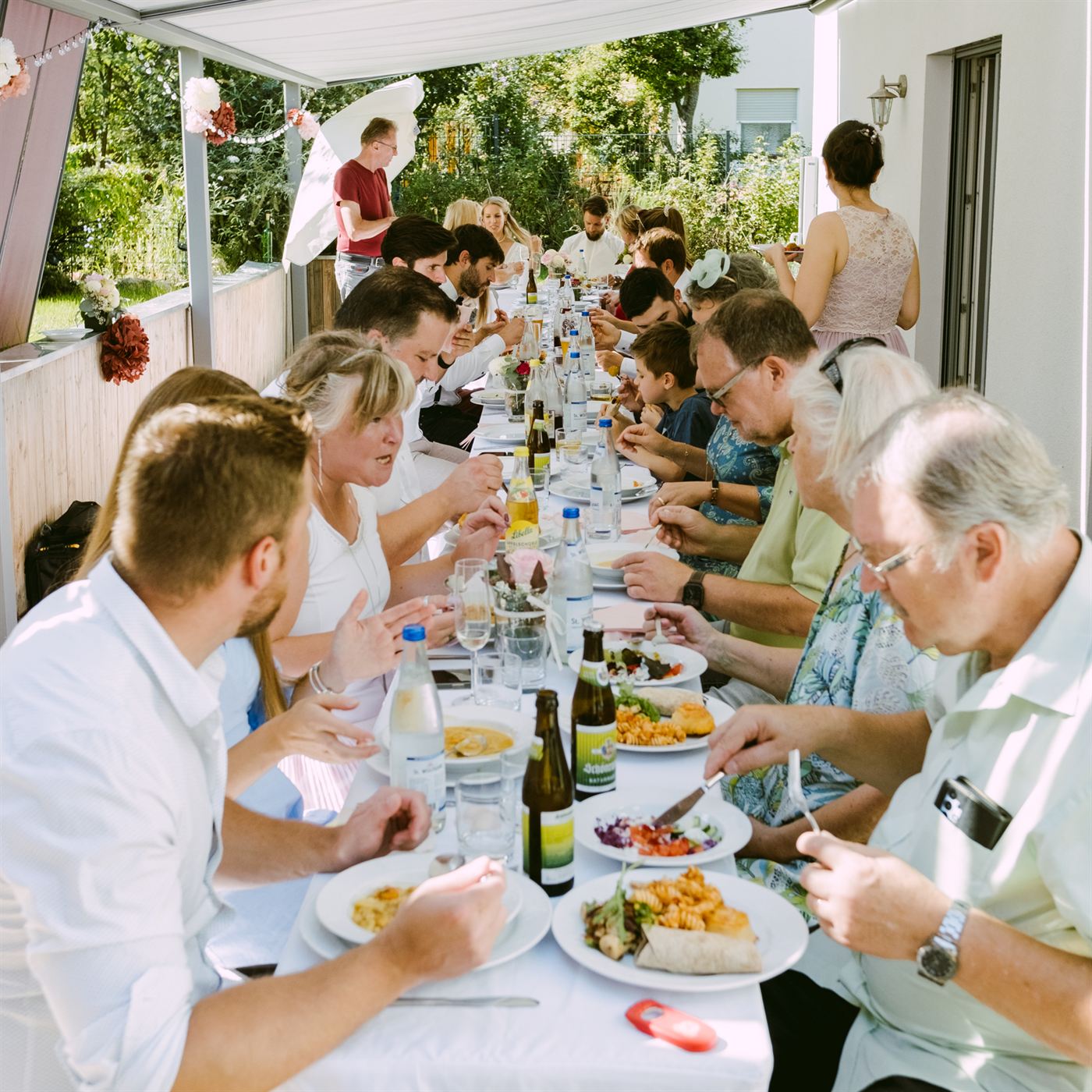 Festlich gedeckte lange Tafel unter einer Markise im Garten, an der Hochzeitsgäste gemeinsam essen.