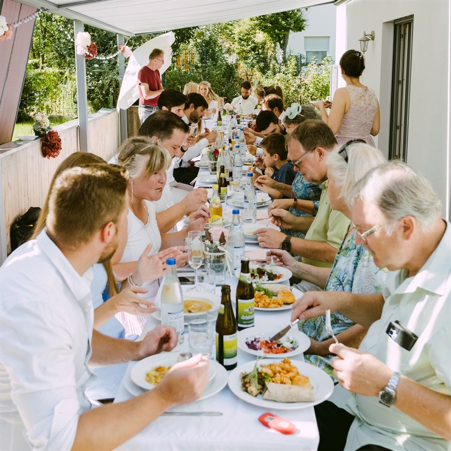 Festlich gedeckte lange Tafel unter einer Markise im Garten, an der Hochzeitsgäste gemeinsam essen.