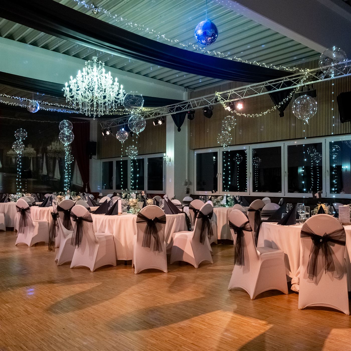 Festlich dekorierter Hochzeitssaal mit Kronleuchter, Discokugel und eingedeckten Tischen mit weißen Stuhlhussen und schwarzen Schleifen.