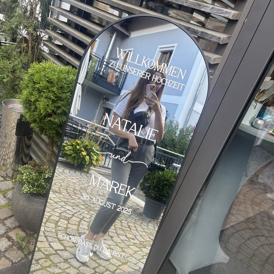 Ovaler Spiegel mit handgeschriebenem Willkommensschild „Willkommen zu unserer Hochzeit – Natalie und Marek – 30. August 2025“ vor einer Hochzeitslocation.