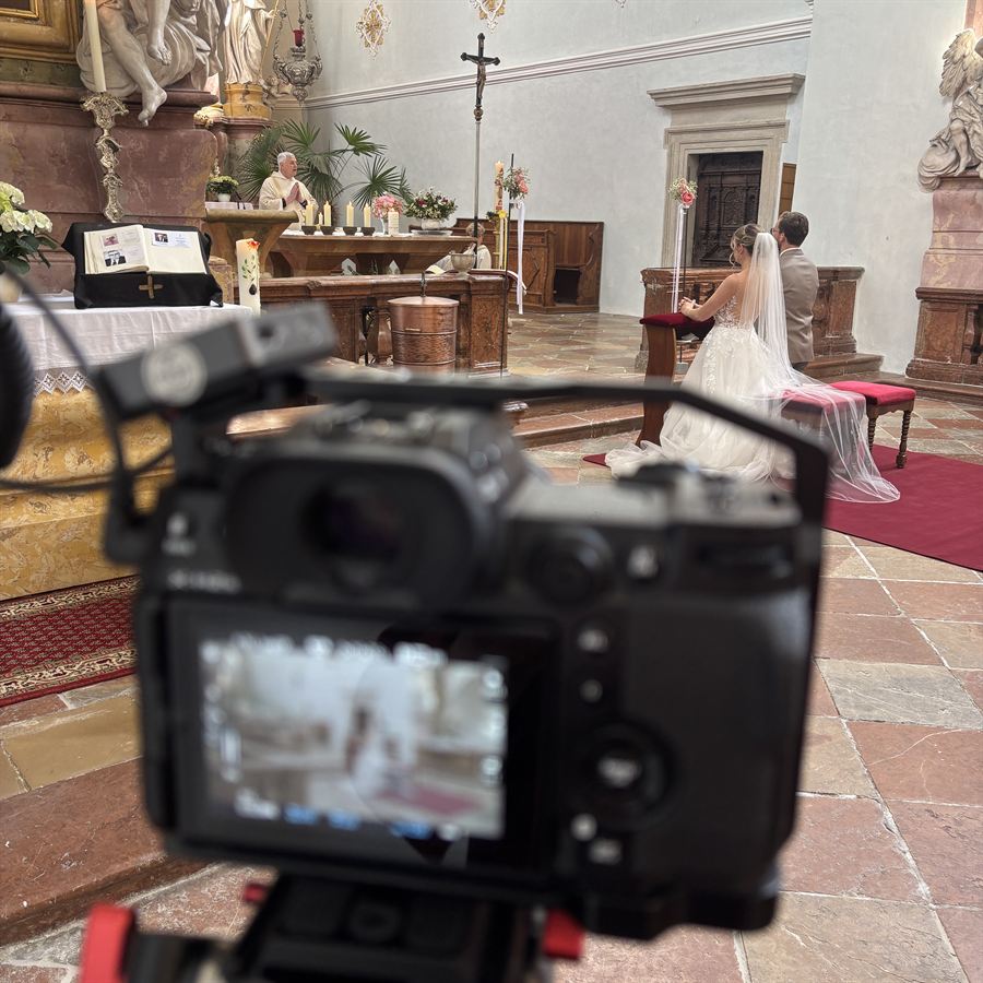 Filmkamera im Vordergrund bei einer kirchlichen Trauung, im Hintergrund kniendes Brautpaar vor dem Altar.